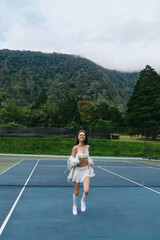 Obraz premium Young woman enjoying a refreshing drink on a tennis court surrounded by lush green mountains, exuding joy and vitality, perfect for healthy lifestyle concepts