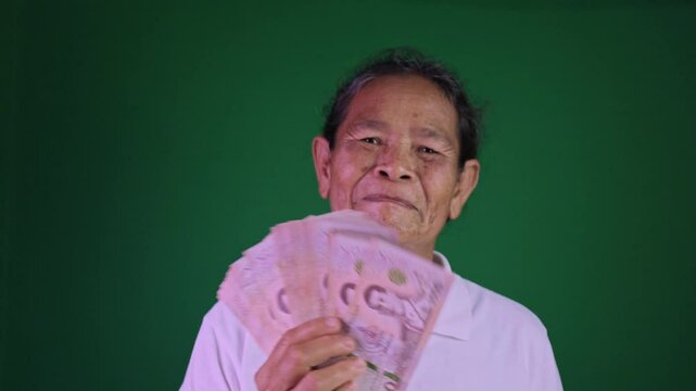 Happy elderly Asian woman holding Thai baht banknotes with big smile, Concept of financial success, luck, and retirement joy on chroma key green screen background, Grandmother happily with a money