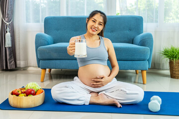 Portrait of pregnant Asian woman wearing a sports bra sitting on a sofa stroking her belly and drinking calcium-fortified milk at living room at home, pregnancy and motherhood concept.