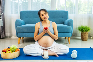 Asian pregnant woman sits on yoga mat on living room floor, hold apple after exercises at home. Beautiful pregnancy mother with big belly eats healthy food and do workout for good health. Happy family