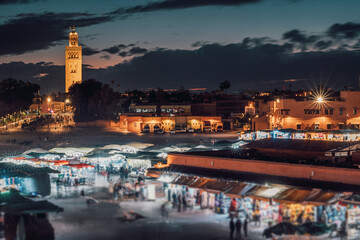 Illuminated stalls and bustling crowds fill Djemaa el Fna market in Marrakech, Morocco, as dusk settles, with the Koutoubia Mosque minaret
