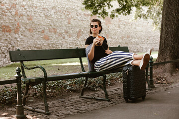 Young woman eating sandwich while relaxing on bench with suitcase