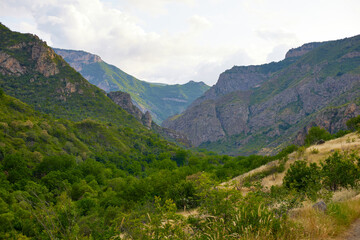 Naklejka premium Scenic view of lush green hills and dramatic red cliffs under a bright blue sky with white clouds, showcasing the beauty of untouched mountainous nature and diverse rocky terrain.