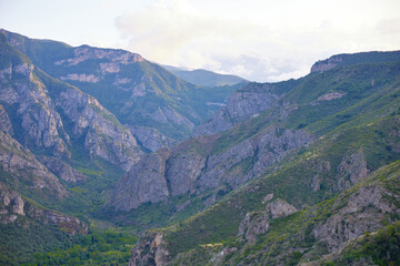 Fototapeta premium Scenic view of lush green hills and dramatic red cliffs under a bright blue sky with white clouds, showcasing the beauty of untouched mountainous nature and diverse rocky terrain.