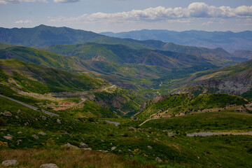 Fototapeta premium Scenic view of lush green hills and dramatic red cliffs under a bright blue sky with white clouds, showcasing the beauty of untouched mountainous nature and diverse rocky terrain.