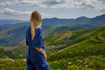 Naklejka premium Woman standing on a mountain peak with arms outstretched, enjoying a breathtaking panoramic view of green hills and valleys under a partly cloudy sky, symbolizing freedom and connection with nature.