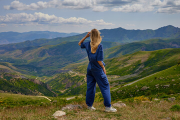 Naklejka premium Woman standing on a mountain peak with arms outstretched, enjoying a breathtaking panoramic view of green hills and valleys under a partly cloudy sky, symbolizing freedom and connection with nature.