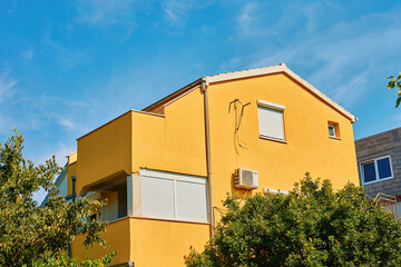 Yellow house with balconies, roller shutters, and an air conditioner against blue sky. Modern suburban building with bright facade