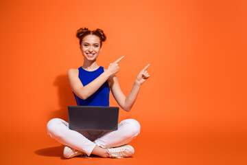 Smiling red-haired young woman pointing upwards while sitting cross-legged with a laptop against an...