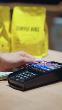 Close-up of person in vintage outfit holding card over a card reader to make payment, with wrist adorned with bracelets and a watch, yellow coffee bags and menu papers are visible on table