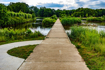 Landscape of Kapermolen Park with pedestrian path over water between Demer River and tributary pond, front view, calm water, and leafy trees around and in background, cloudy day in Hasselt, Belgium