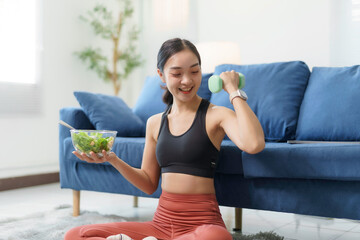 Happy asian sportswoman lifting light dumbbell and holding bowl of healthy salad while sitting on floor in living room at home