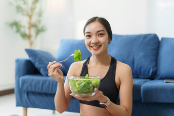 Happy Asian girl enjoying a fresh bowl of salad after completing fitness exercises at home, embracing a healthy and active lifestyle