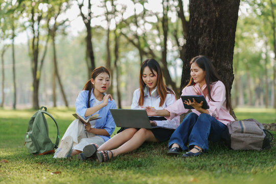 Three asian female students sitting on grass under a tree, using laptop and tablet, working together on a project