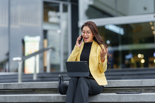 Asian businesswoman working remotely using tablet and mobile phone sitting on stairs outside office building - Powered by Adobe