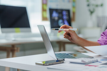 Businesswoman holding a pen and pointing at laptop screen while working with financial documents in...
