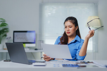 Focused businesswoman reading documents and working with laptop at office desk, holding eyeglasses in hand