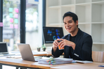 Young entrepreneur smiling while holding a coffee cup, working diligently at his modern desk with a laptop and documents nearby