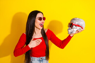 Young woman wearing red shirt and sunglasses holding a decorated skull against a vibrant yellow...