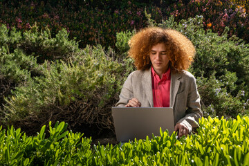 A young executive dressed casually with red afro hair. He is working on his laptop in the middle of a public park surrounded by plants. Sustainability and technology