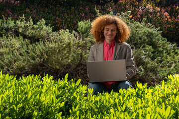 A young executive dressed casually with red afro hair smiles while working. He is working on his laptop in the middle of a public park surrounded by plants. Sustainability and technology