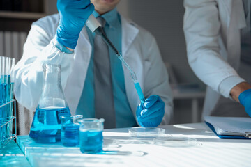 Two scientists using pipette and petri dish doing research with blue liquid reagents in laboratory...