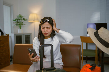 Young woman sitting on sofa with packed suitcase and holding her head while looking at mobile...