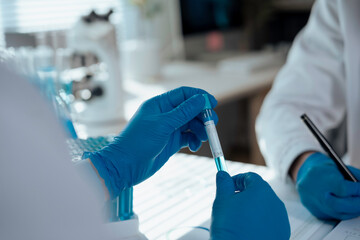 Two scientists wearing blue gloves are examining a test tube containing blue liquid in a laboratory setting, with a microscope visible in the background