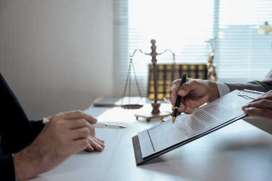 Lawyer holding contract and showing client where to sign with pen, with scales of justice in background