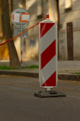 In a busy urban area, a bright red and white traffic barrier blocks a street. A nearby sign warns drivers of road work and suggests alternative routes around the blockage