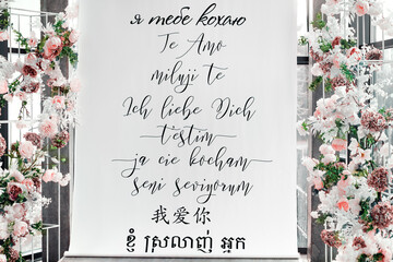 Photo of a real wedding venue with floral decorations and a romantic backdrop featuring “I love you” written in multiple languages. Text is part of the actual scene, not digitally added.