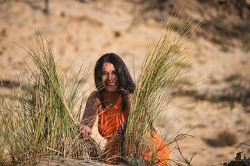 A young, beautiful brunette woman in an orange dress is kneeling and leaning between the reeds...