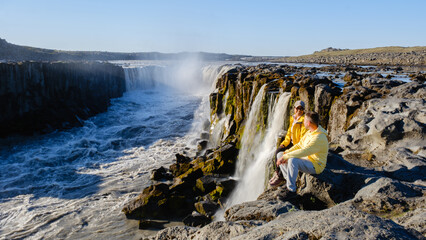 Visitors sit on rocky terrain, captivated by the mighty Dettifoss waterfall in Iceland.
