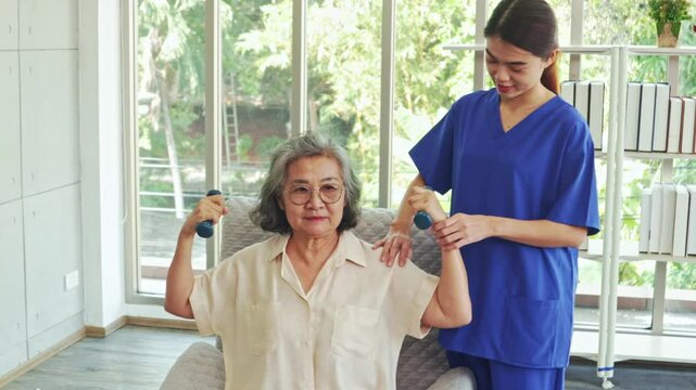 Female physiotherapist is providing nursing care an elderly female patient with muscular dystrophy training her to do dumbbell exercises rehabilitate her muscles and prevent beriberi.