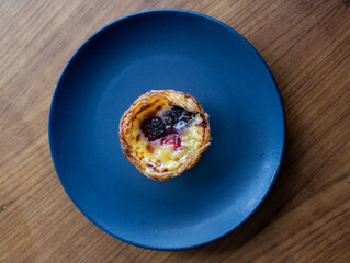 A close-up of a traditional Portuguese custard tart (Pastel de Nata) served on a dark blue plate over a wooden table. The tart is topped with fresh berries, including blackberry and raspberry.