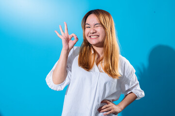 Fototapeta premium Asian woman with long straight hair in a white shirt smiles brightly while making an OK hand gesture with one hand and winking playfully against a plain blue background in studio lighting