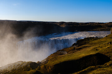 Water plunges powerfully over rocky edges at Dettifss Iceland, creating a misty atmosphere amidst lush greenery.
