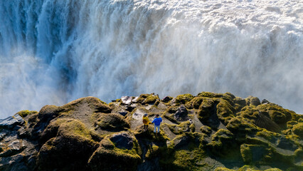 Adventurers stand near a powerful Dettifoss waterfall in Iceland, surrounded by lush green moss and rocky terrain.
