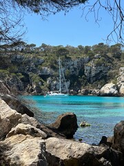 A white sailboat anchored in a turquoise bay on Menorca, framed by rocky cliffs and pine trees under a clear blue sky, showcasing the island’s natural beauty and tranquility.
