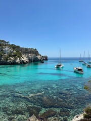 Sailboats anchored in the crystal-clear turquoise waters of Cala Macarella, Menorca, surrounded by limestone cliffs and lush Mediterranean vegetation on a sunny summer day.
