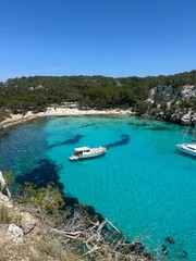 Crystal-clear turquoise waters of Cala Macarella beach in Menorca, surrounded by pine trees and rocky cliffs, with boats anchored in the peaceful bay.
