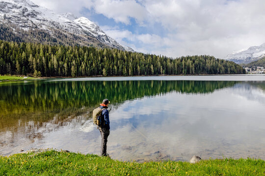 A hiker stands by a serene lake, reflecting the surrounding mountains and lush forest.
