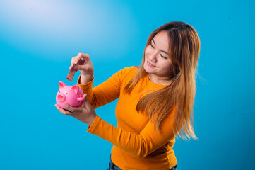 Asian woman in orange shirt smiling while putting a coin into a cute pink piggy bank, standing against a bright blue background, symbolizing savings and smart financial habits