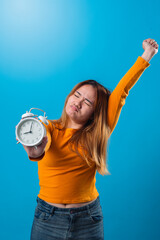 Asian woman stretching with one arm up and holding a white alarm clock forward in the other hand, showing a sleepy expression in front of a solid blue background