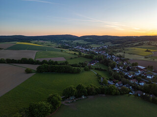 Drone panorama of Brombachtal, Germany at dusk, showing peaceful rural homes, farmlands, forests, and gentle hills.

