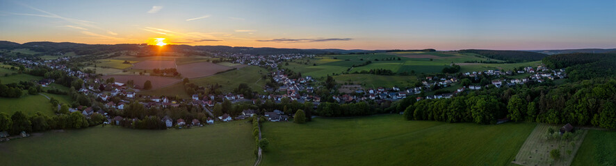 Aerial panorama of Brombachtal, Germany at sunset with rolling hills, green fields, forest, and a peaceful countryside village.
