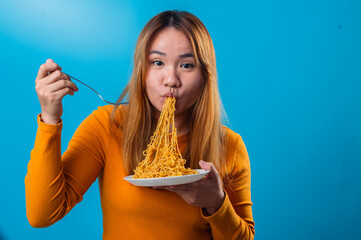 Asian woman in orange shirt eagerly slurping a forkful of instant noodles with eyes closed, showing...