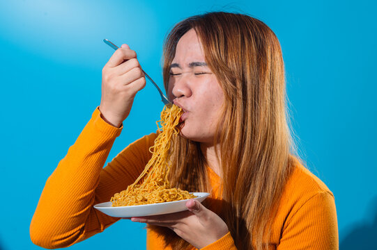 Asian woman in orange shirt eagerly slurping a forkful of instant noodles with eyes closed, showing enjoyment and hunger, in front of a clean blue background