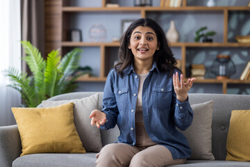Young Indian girl in a denim shirt sitting on the couch at home and talking to the camera while gesturing with her hands