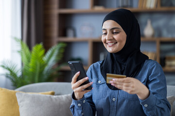 Smiling young Muslim woman wearing hijab sitting on sofa at home and using mobile phone and credit...
